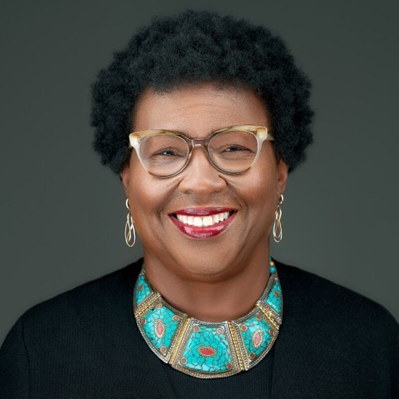 The image shows a smiling Black woman with short, curly hair and glasses. She is wearing a black top and a turquoise and coral necklace. Her earrings are gold. The background is a solid, muted gray. She appears to be in a studio setting.